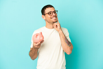 Brazilian man holding a piggybank over isolated blue background thinking an idea while looking up
