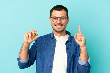 Brazilian man holding a Bitcoin over isolated blue background showing and lifting a finger in sign of the best