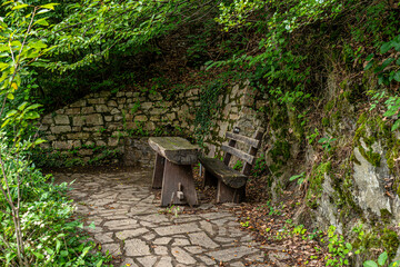 Fototapeta na wymiar An old bench and a wooden table standing on a stone floor next to a water spring in the forest, a stone wall visible.