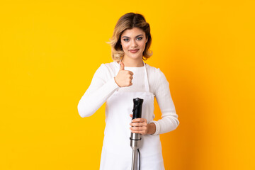 Girl using hand blender isolated on yellow background giving a thumbs up gesture