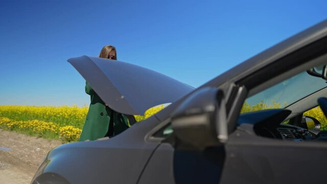 Woman in the coat opens the hood of the car.