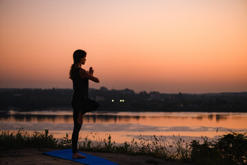 Fototapeta premium Morning sunrise at water and city horizon with healthy woman standing in balance yoga Vrksasana tree position