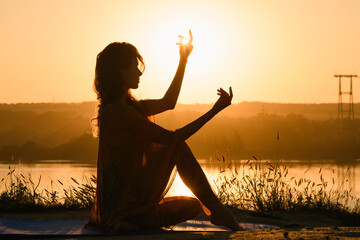 Young woman silhouette sitting against sun in warm morning sun light, meditation yoga fitness practice outdoors