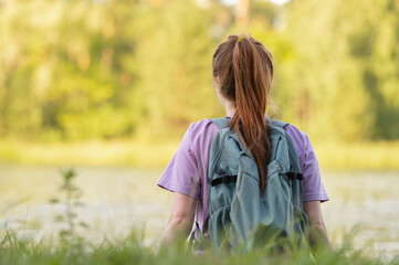 Redhead girl sits on the grass near the water in a beautiful summer forest. Young woman travels with backpack