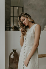 Fototapeta premium Pretty elegant blonde curly woman in linen beige dress smiles and looks into camera. Young lady with massive white earrings poses in cozy bathroom.