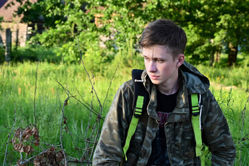 young tired worker in protective overalls is resting, sitting outdoors against background of green...