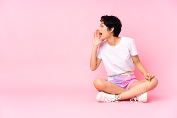 Young Vietnamese woman with short hair sitting on the floor over isolated pink background shouting with mouth wide open to the lateral