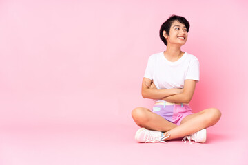 Young Vietnamese woman with short hair sitting on the floor over isolated pink background with arms crossed and happy