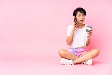 Young Vietnamese woman with short hair sitting on the floor over isolated pink background holding coffee to take away and a mobile