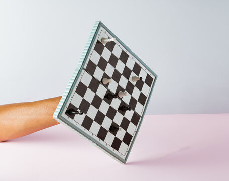 Hand holding white and black chess board with figures on pink and grey background. Minimal concept.