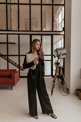Self-confident young calm woman in stylish black silk home suit looks into window, stands in cozy living room and holds laptop and newspaper.