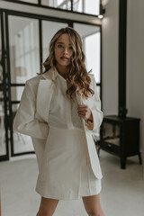 Cheerful stylish curly blonde woman in trendy white skirt suit and blouse looks into camera and poses in beautiful living room with fireplace.