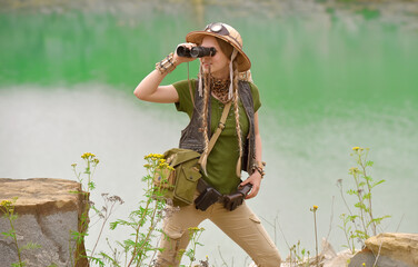 A young girl is dressed up as an explorer. She 
is seen in a green lake environment observing 
the...