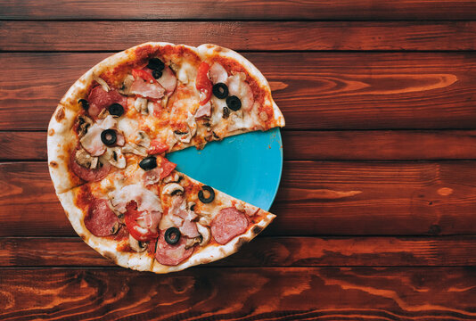 Freshly baked pizza with sausage, bacon, mushrooms, tomatoes and cheese lies on a blue plate and a brown wooden background without one slice. Fast food concept. View from above.