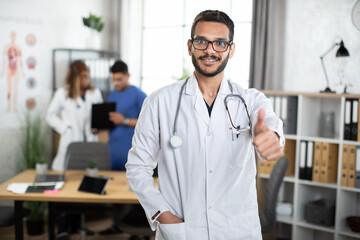 Portrait of 30-aged Indian Asian male doctor in a clinic room, showing thumb up and smiling to camera, with two busy colleagues in the background. Copy space