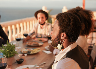 Multiracial young people enjoy dinner at home on patio while wearing safety face mask under chin