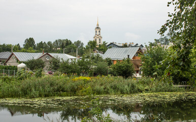 Fototapeta na wymiar panoramic view of the old town with historical wooden houses and an Orthodox church among the green foliage of trees on the river bank with a reflection on a cloudy day in Suzdal Russia