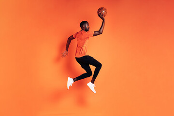 Fototapeta na wymiar Male athlete jumping with basket ball. Side view of sportsman exercising over orange background.