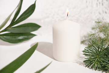 Obraz na płótnie Canvas white candle and green leaves on white table with textured background. burning candle, minimal style home decor concept