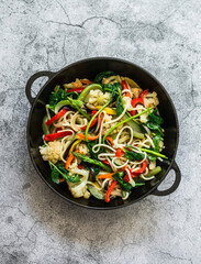 Vegetarian noodles with vegetables - cauliflower, zucchini, pepper, spinach, eggplant, carrot wok in a frying pan on a gray background, top view