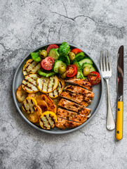 Barbecue plate - chicken breast, yellow zucchini, grilled potatoes and fresh vegetable salad with pesto sauce dressing on a grey background, top view
