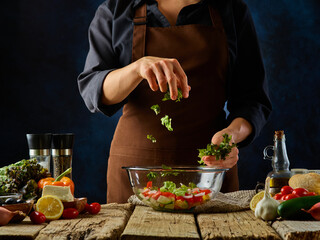 The chef mixes the ingredients for the Greek salad. He tosses greens into a large glass bowl. Ingredients for Greek salad. Bright paints, dark blue background. Hotel, restaurant, fitness bar.