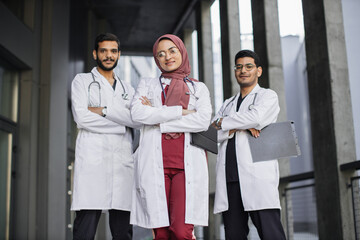 Front view of high-skilled team of three young confident Saudi Arab doctors, standing outside the...