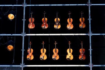 Ten violins or violas hanging in a metal frame on stage during a performance with two stage lights on the left
