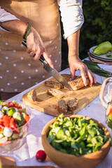 Cutting wholegrain bread on table. Preparing fresh salad with baguette for garden party. Healthy eating
