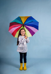 a happy little girl in a raincoat and rubber boots stands and holds a multi-colored umbrella on a blue background with a copy of the space