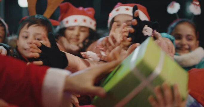 4K Santa Claus gives wrapped gift boxes to cute little children . Two Santa clauses helping each other to give gifts to queue of children . Many kids standing and waiting in line queue for a gift .