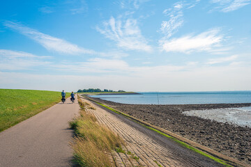 A man and a woman are cycling on a path at the bottom of a Dutch dike and along the Oosterschelde. The dike is reinforced with stones and partly covered with green algae. It is summertime now.