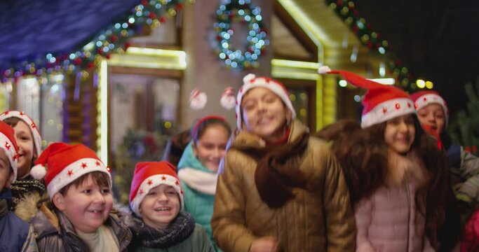 Happy children are jumping , laughing  and looking camera into front of the beautiful house decorated with lights and garlands.  Portrait of many kids with Santa hat and costumes at Christmas night .