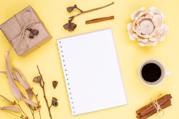 Blank diary with spring, gift in kraft paper, candlestick, autumn dry eucalyptus leaves and seeds, a cup of coffee on a yellow background. Flat lay, copy space