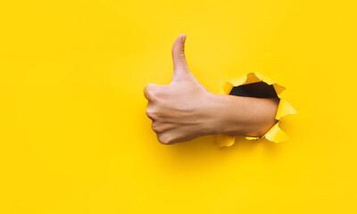 The female hand shows a thumb up gesture (like). Torn hole in yellow paper. The concept of positive...
