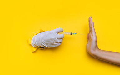 The doctor hand in a white latex glove holds a syringe for vaccination, but collides with a hand in the form of a Stop gesture. Coronavirus anti-vaccination concept.Yellow paper, torn hole, copy space