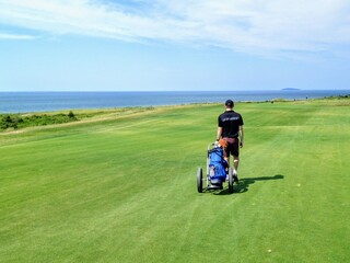 A young male walking his golf bag using a golf cart walking down a large golf fairway with the...