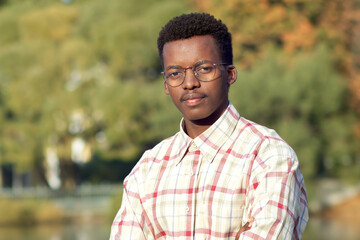 Naklejka na ściany i meble Portrait of serious handsome black African Afro American ethnic guy, young man in glasses standing in golden autumn, fall park with his hands, arms crossed, looking at camera in shirt near lake