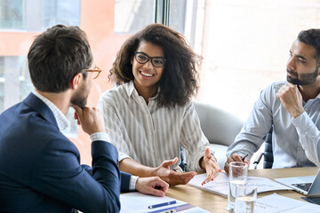 Smiling female manager talking to businessmen team discussing financial sales research plan at boardroom meeting table. Multiethnic team working together developing business strategy in modern office. - obrazy, fototapety, plakaty