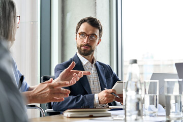 Serious Caucasian bearded male ceo wearing glasses looking at female manager controlling discussing corporation project report at table. Executive managers leaders working together in modern office. - obrazy, fototapety, plakaty