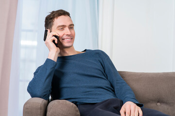 Portrait of happy positive guy, young cheerful man is chatting, talking on cell mobile phone, having good nice conversation on smartphone, sitting on couch or sofa in living room at home, smiling