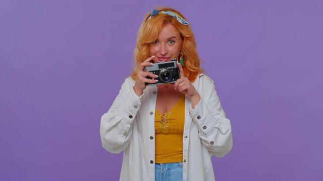 Red hair stylish girl tourist in white shirt. Photographer taking photos on retro camera and smiling. Young woman on purple studio background. People sincere emotions. Travel, summer holiday vacation