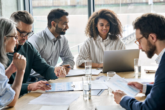 Diverse executive businesspeople discuss corporation financial plan at boardroom meeting table. Multiracial team negotiate project developing business strategy doing paperwork using laptops in office.