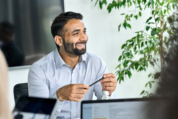 Friendly happy smiling ceo Indian businessman holding pen in hand looking listening to colleague discussing global corporation research report at table. Executive manager working in modern office.