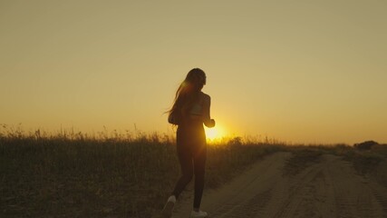 Fototapeta na wymiar A girl in headphones runs along the road at sunset, a young woman makes a morning jog, a healthy lifestyle outdoors, a long-distance race, preparation for victory in a sports competition