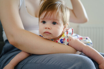 Fototapeta premium Small caucasian baby girl in the lap of her mother while sitting on the sofa at home - motherhood and parenting concept real people growing up in bright summer day