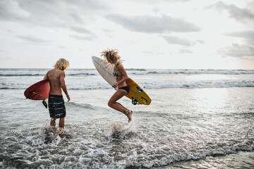 Slender guy and girl jump in sea water and hold surfboards