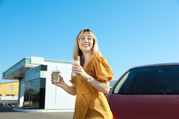 Beautiful young woman with coffee and hot dog near car at gas station