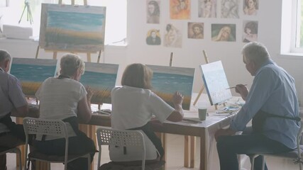 A young female art teacher demonstrates the technique of painting with acrylic paints on canvas for a group of retirees. Group of senior men and women