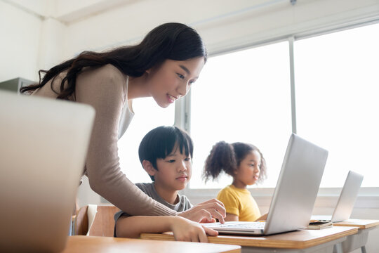 Asian female teacher teaching Asian boy and African American girl learning on laptop computers at international primary school